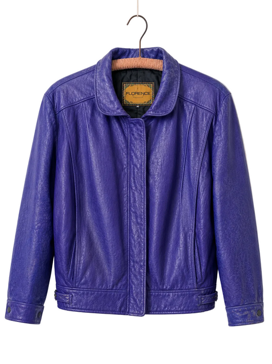 A vibrant purple genuine leather jacket hanging on a wooden hanger against a plain white wall. The jacket features a pointed collar, a zip front closure, and angled hand pockets.